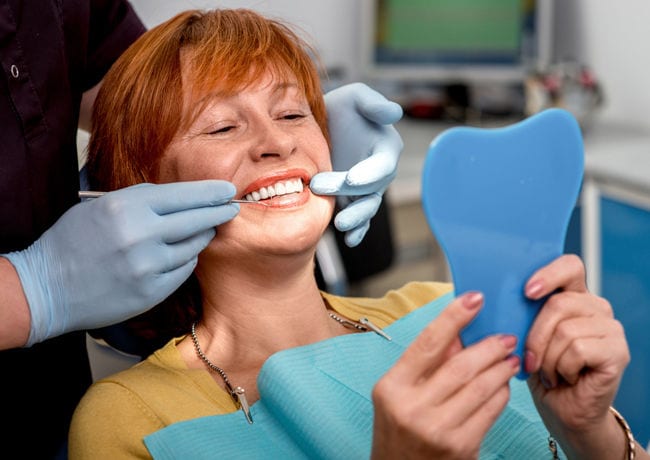 A woman patient receives dental work and smiles while looking into a hand mirror to see the color and fit of her new dentures.